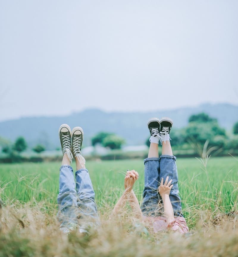 jambes tendues de deux adolescents allongés dans une prairie, se tenant la main. Atmosphère d'insouciance, de complicité et de légèreté, symbolisant les bienfaits de la sophrologie pour les adolescents.