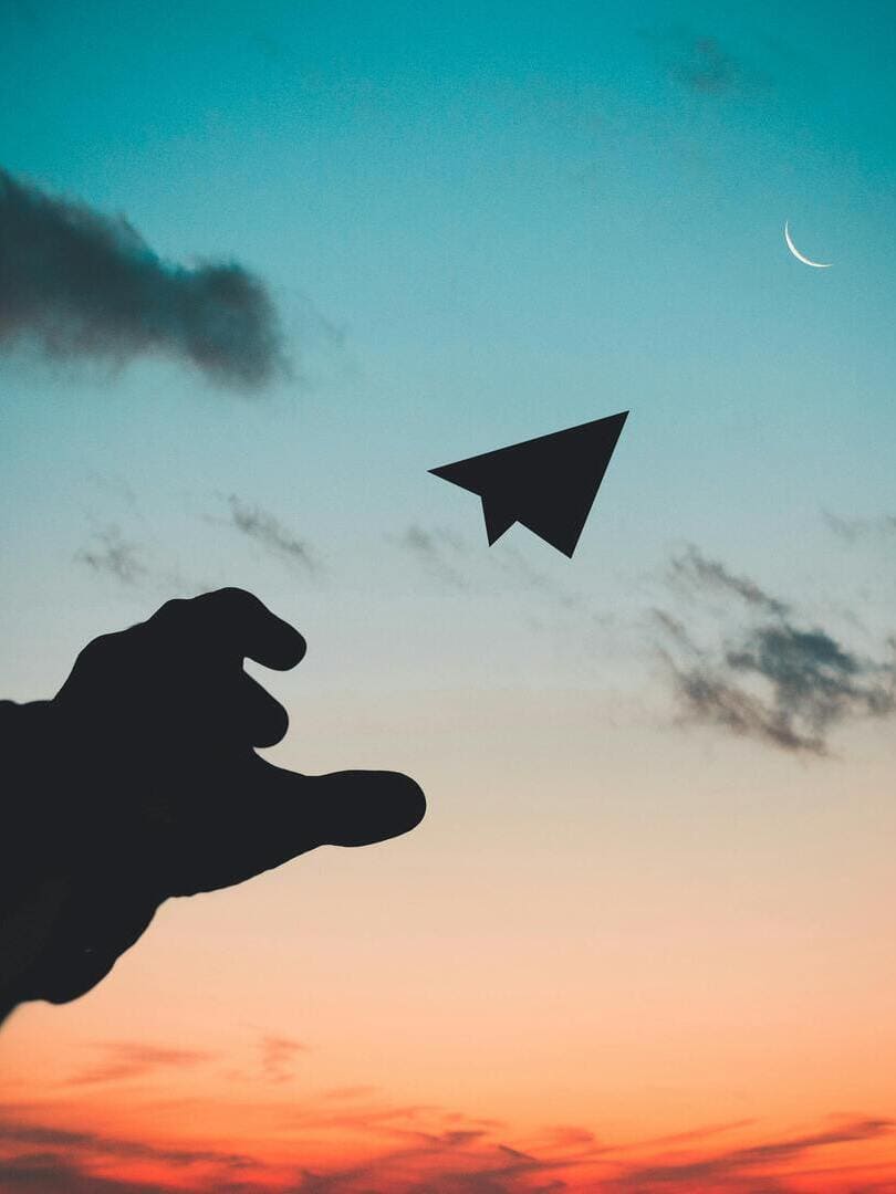 Silhouette d'une main lançant un avion en papier vers la lune, dans un ciel aux nuances douces de bleu et d'orange. Atmosphère paisible, symbolisant la créativité, l'espoir et la sérénité