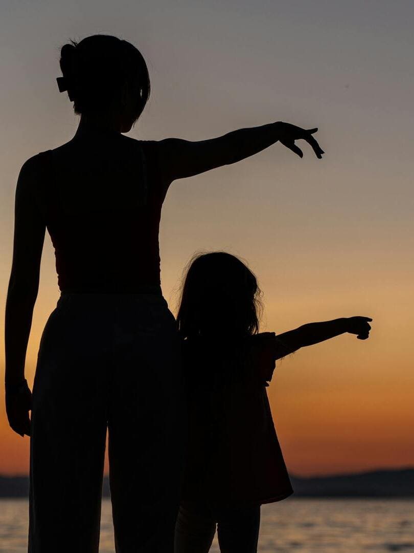 Silhouettes d'une mère et de sa fille regardant au loin, pointant dans le même direction sous un coucher de soleil aux tonalités apaisantes. Symbolise la complicité, le lien mère fille, et la projection vers l'avenir serein et confiant.