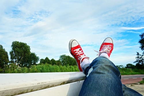 Jambes croisées chaussées de baskets rouges, posées de façon décontractées sur un banc avec en arrière-plan un ciel bleu et de la verdure. Illustration du bien-être, de la détente et de l'amélioration de la qualité de sommeil et de repos grâce à la sophrologie